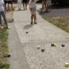 Des personnes jouent au jeu de boules carrées en plein air, lançant des cubes de bois colorés - principalement jaunes et gris - sur un chemin de gravier, tandis que d'autres observent ou se préparent à rejoindre le jeu.