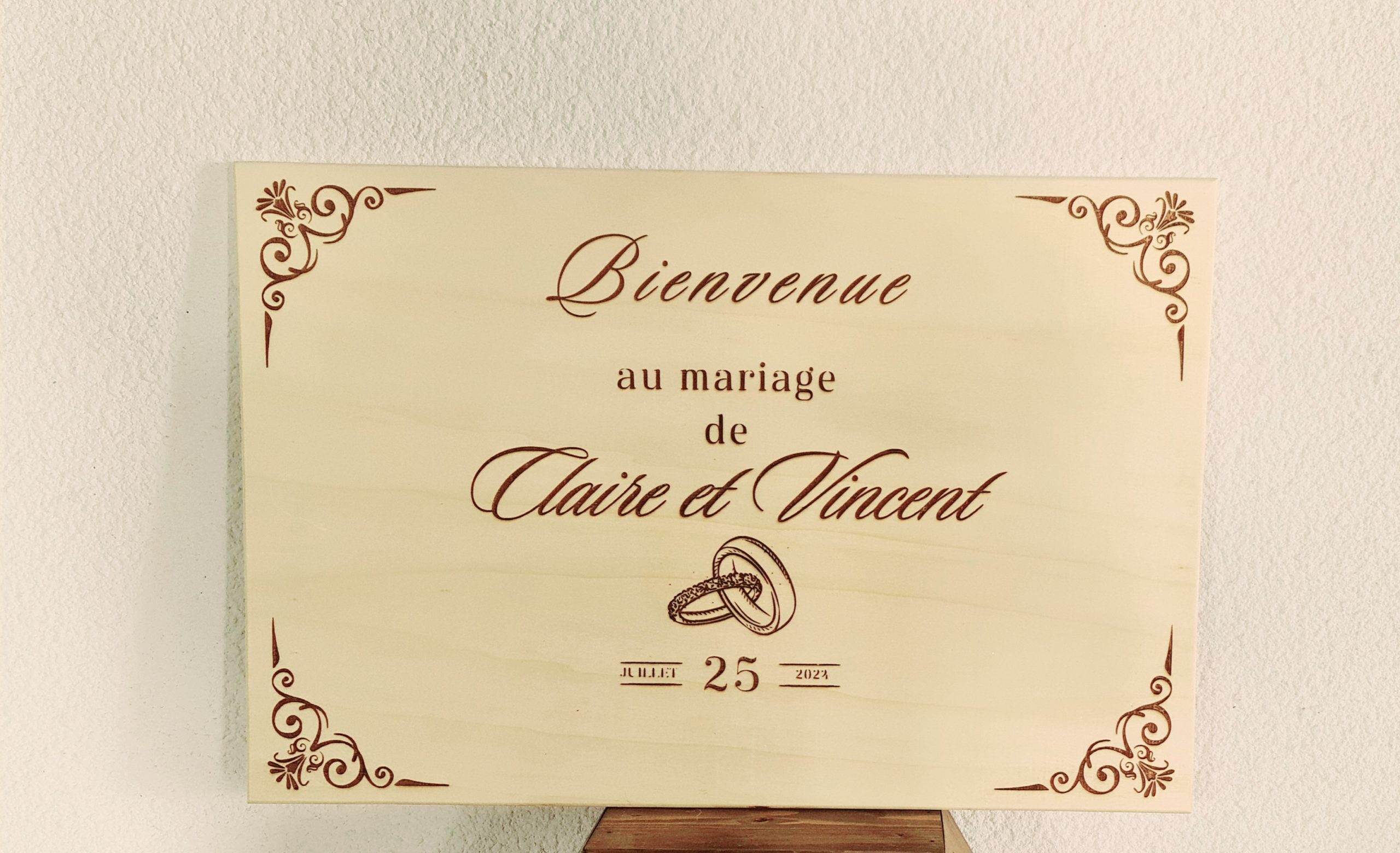 Un panneau en bois aux coins ornés indique : Bienvenue au mariage de Claire et Vincent avec des alliances entrelacées et la date du 12.08.2023 en dessous, souhaitant la bienvenue aux invités au mariage.