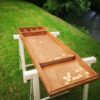 Une table de billard-hollandais avec des palets est installée sur une pelouse au bord d'une rivière, entourée d'arbres et de buissons verts.