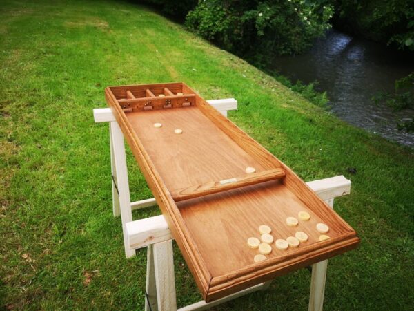 Une table de billard-hollandais avec des palets est installée sur une pelouse au bord d'une rivière, entourée d'arbres et de buissons verts.