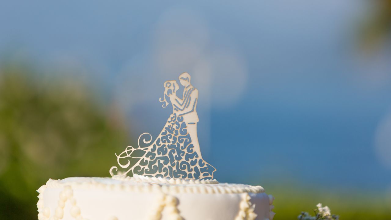 Un gâteau de mariage blanc surmonté d'une figurine de couple ajouré, représentant des mariés enlacés, se dresse devant un arrière-plan extérieur flou de verdure et de ciel bleu.