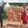 Puissance 4 Deux adultes en tenue de soirée jouent à Puissance 4 XXL sur l'herbe. La femme tient un verre tandis que l'homme place un disque, avec des buissons et des arbres en arrière-plan.