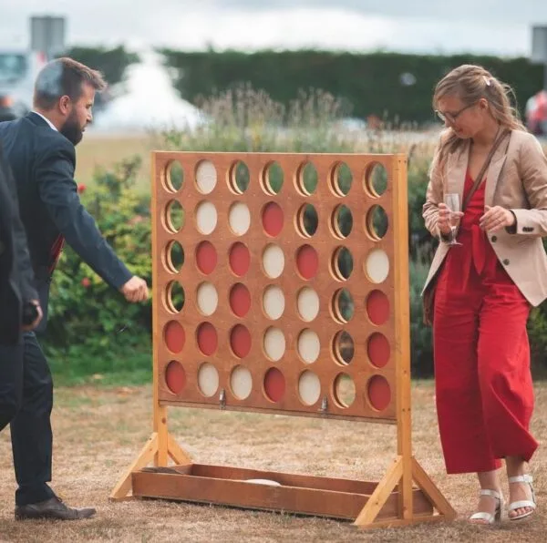 Deux adultes en tenue de soirée jouent à Puissance 4 XXL sur l'herbe. La femme tient un verre tandis que l'homme place un disque, avec des buissons et des arbres en arrière-plan.
