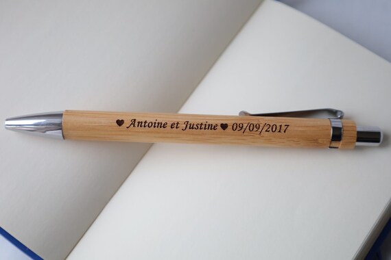 Un stylo en bois aux accents métalliques gravés Antoine et Justine ♥ 09/09/2017 repose sur les pages ouvertes d'un carnet vierge.