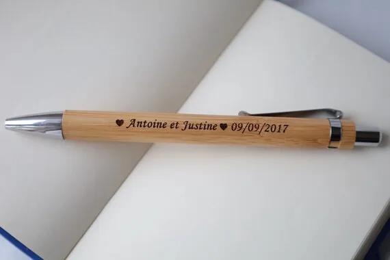 Un stylo en bois aux accents métalliques gravés Antoine et Justine ♥ 09/09/2017 repose sur les pages ouvertes d'un carnet vierge.
