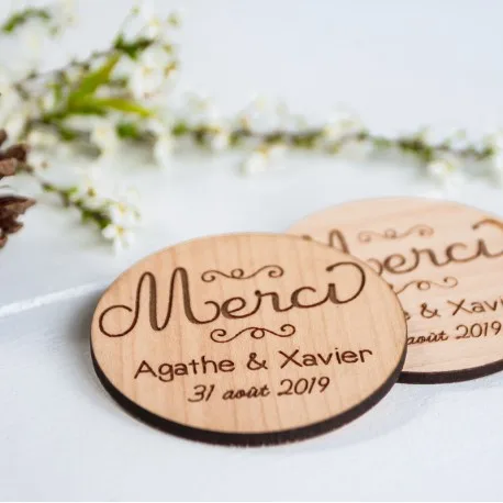 Deux sous-verres ronds en bois gravés de Merci, Agathe & Xavier, et 31 août 2019, posés sur une surface blanche à proximité de petites fleurs blanches.