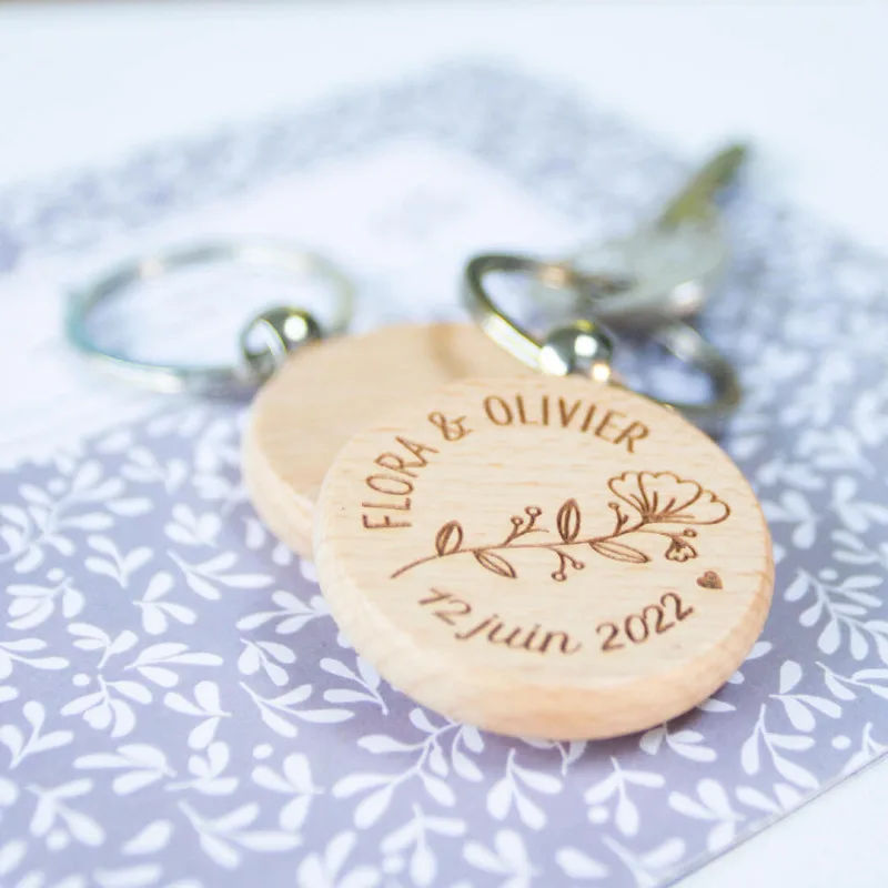 Un porte-clés rond en bois gravé de Flora & Olivier, un motif floral, et du 12 juin 2022. Le porte-clés est attaché aux clés et repose sur une surface à motifs violets et blancs.