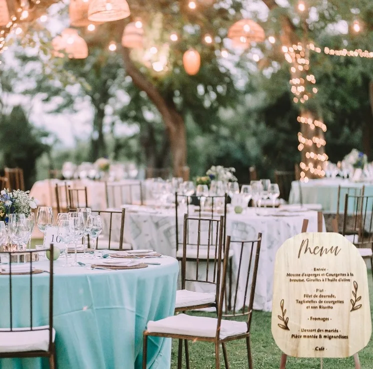 Réception de mariage en plein air avec tables rondes recouvertes de nappes bleu clair, décorées de verres et de fleurs. Guirlandes lumineuses et lanternes sont suspendues au plafond, et un menu de mariage est visible au premier plan. Arbres et verdure entourent la scène.