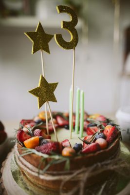 Un gâteau garni de fruits, décoré de deux étoiles dorées scintillantes et d'une décoration dorée scintillante en forme de chiffre 3, ainsi que de trois bougies vertes, repose sur une surface en bois.