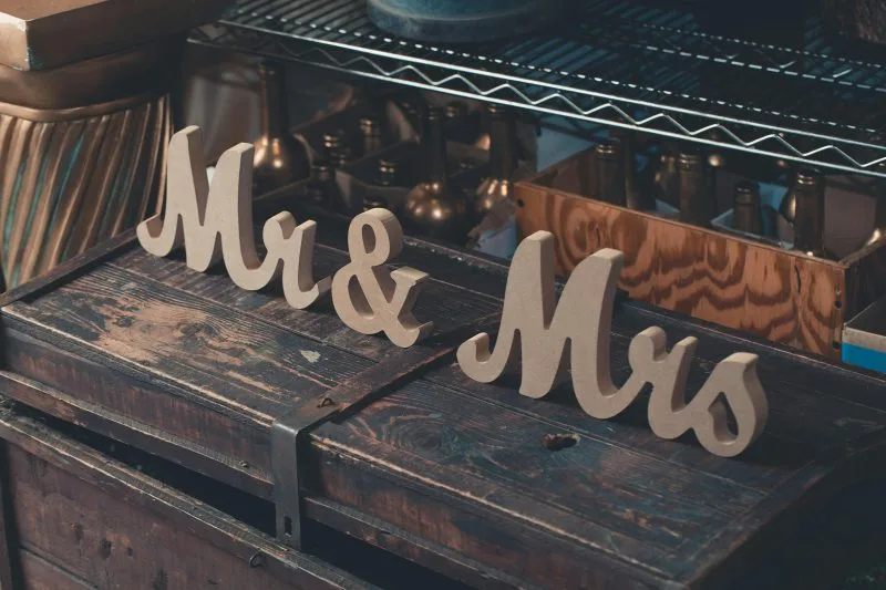 Des panneaux découpés en bois indiquant « M. et Mme » sont placés sur un coffre en bois rustique, ajoutant une charmante touche de décoration pour mariage, avec des étagères et divers objets métalliques en arrière-plan.